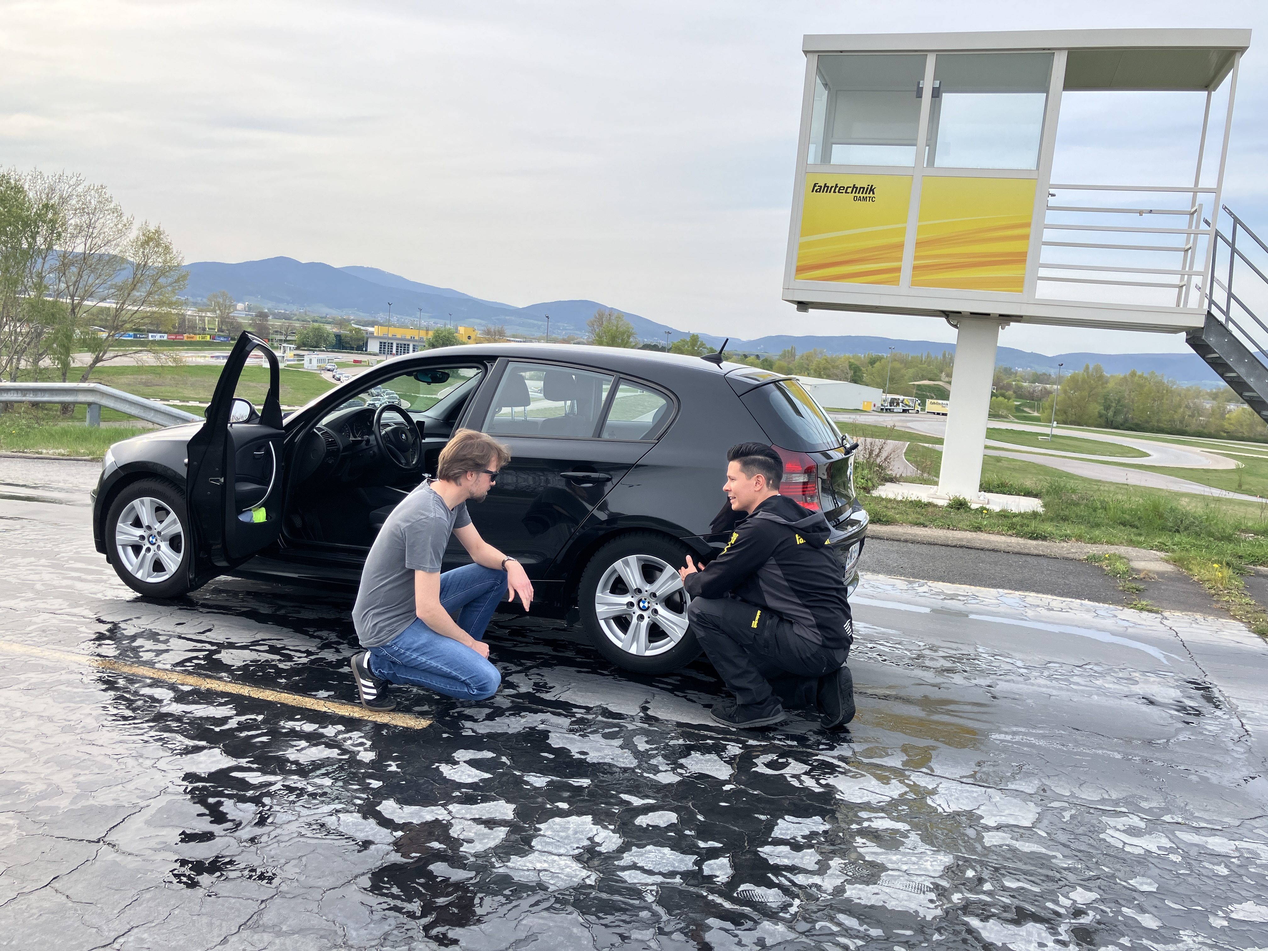 Auf dem Bild sieht man zwei Männer, die vor dem Hinterreifen eines schwaren Fahrzeugs knien. Die Fahrertür des Autos ist geöffnet. Der rechte Mann erklärt dem linken etwas. Die Fahrbahn ist feutcht. Im Hintergrund sieht man eine erhöhte Metallkabine auf der Fahrtechnik steht.