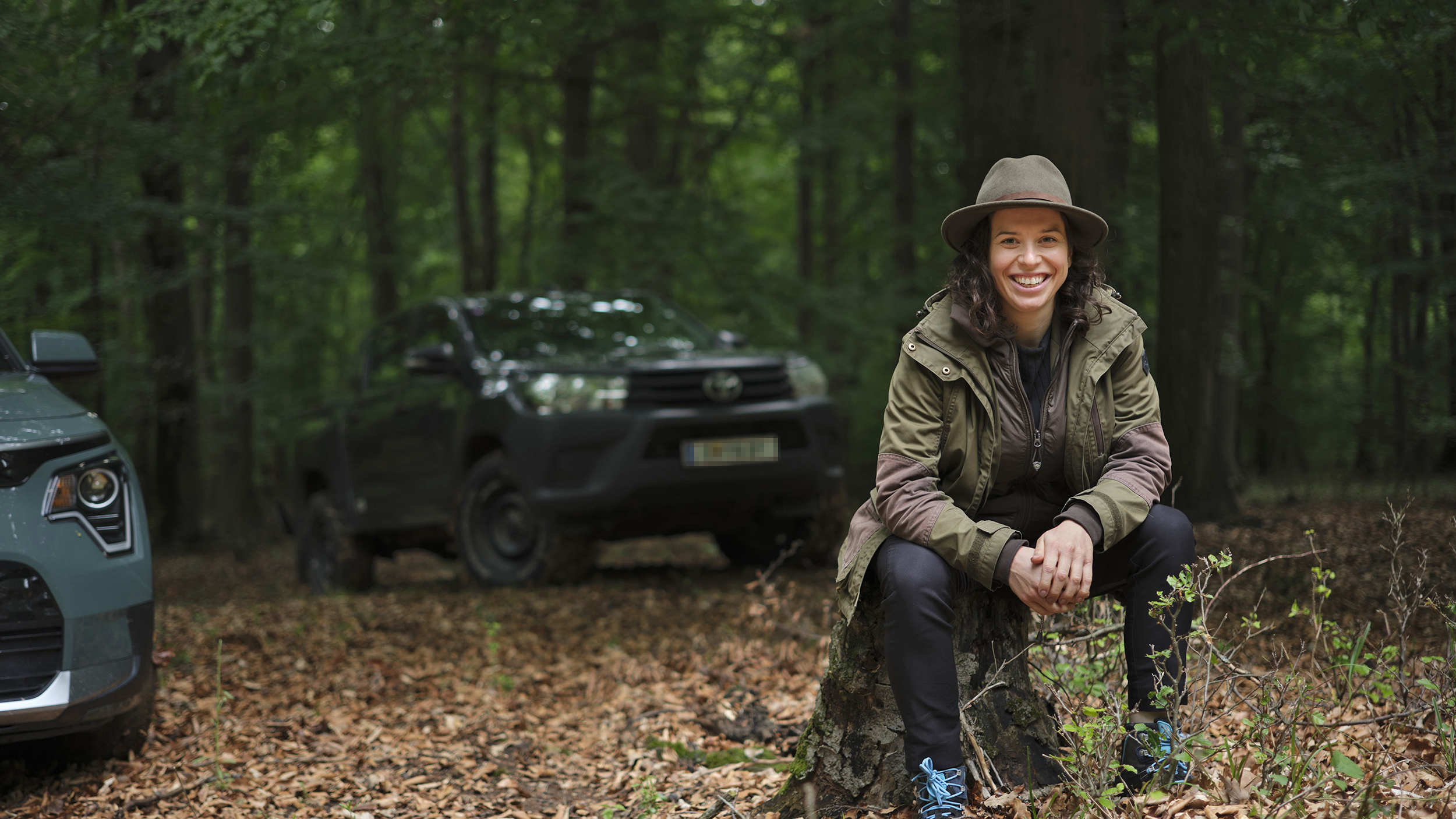 Försterin Stefanie Nievoll sitzt auf einem Baumstamm im Wald. Im Hintergrund ein Toyota Hilux und Bäume.