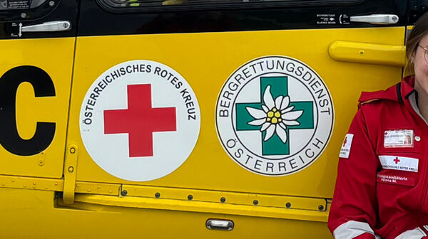 Eine junge Frau in Rotkreuz-Uniform und ein junger Mann in der Uniform der ÖAMTC-Flugrettung sitzen vor einem gelben Hintergrund. Auf diesem erkennt man den Schriftzug ÖAMTC, Christophorus, sowie die Logos der Bergrettung und des Roten Kreuzes.