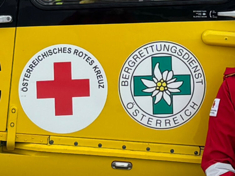 Eine junge Frau in Rotkreuz-Uniform und ein junger Mann in der Uniform der ÖAMTC-Flugrettung sitzen vor einem gelben Hintergrund. Auf diesem erkennt man den Schriftzug ÖAMTC, Christophorus, sowie die Logos der Bergrettung und des Roten Kreuzes.