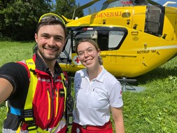 Eine junge Frau in Rotkreuz-Uniform und ein junger Mann in der Uniform der ÖAMTC-Flugrettung stehen vor einem ÖAMTC Notarzthubschrauber.