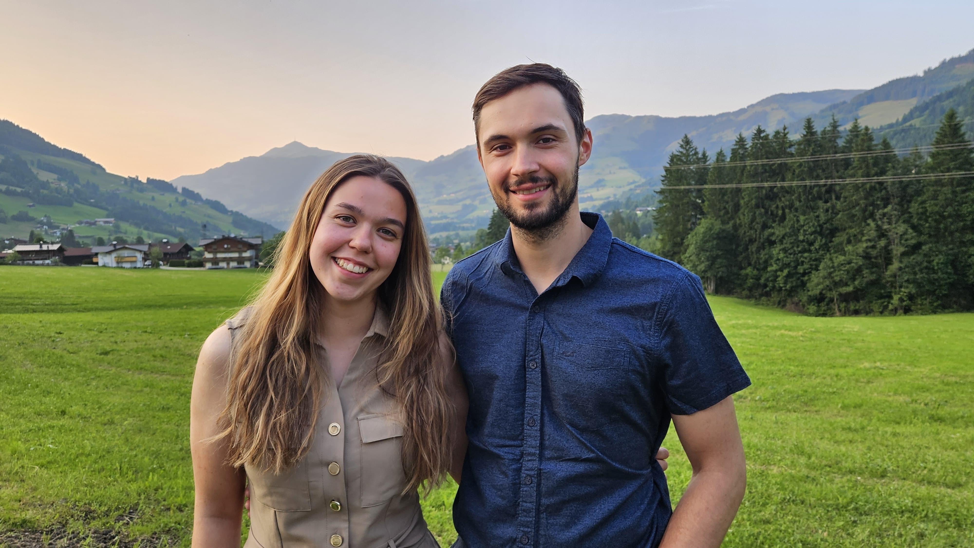 Eine junge Frau in beiger Bluse und ein junger Mann im blauen Hemd stehen auf einer grünen Wiese, im Hintergrund kann man Berge erkennen.