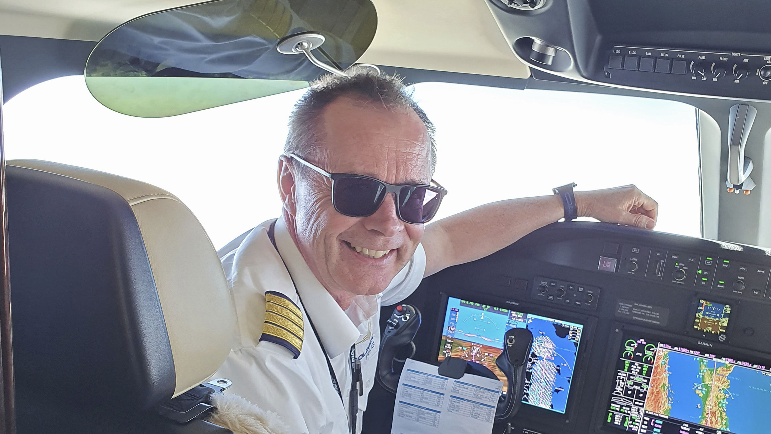 Flugkapitän Harry Gruber sitzt im Cockpit eines Flugzeugs und lächelt. Er hat kurze graue Haare und eine schwarze Sonnenbrille. Er trägt eine Pilotenuniform.