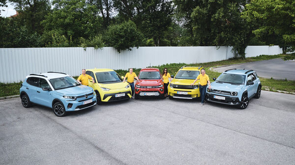 Fünf kleine Elektroautos nebeneinander aufgereiht, stehend von vorne, mit vier Personen