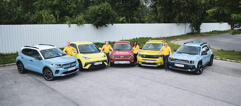 Fünf kleine Elektroautos nebeneinander aufgereiht, stehend von vorne, mit vier Personen