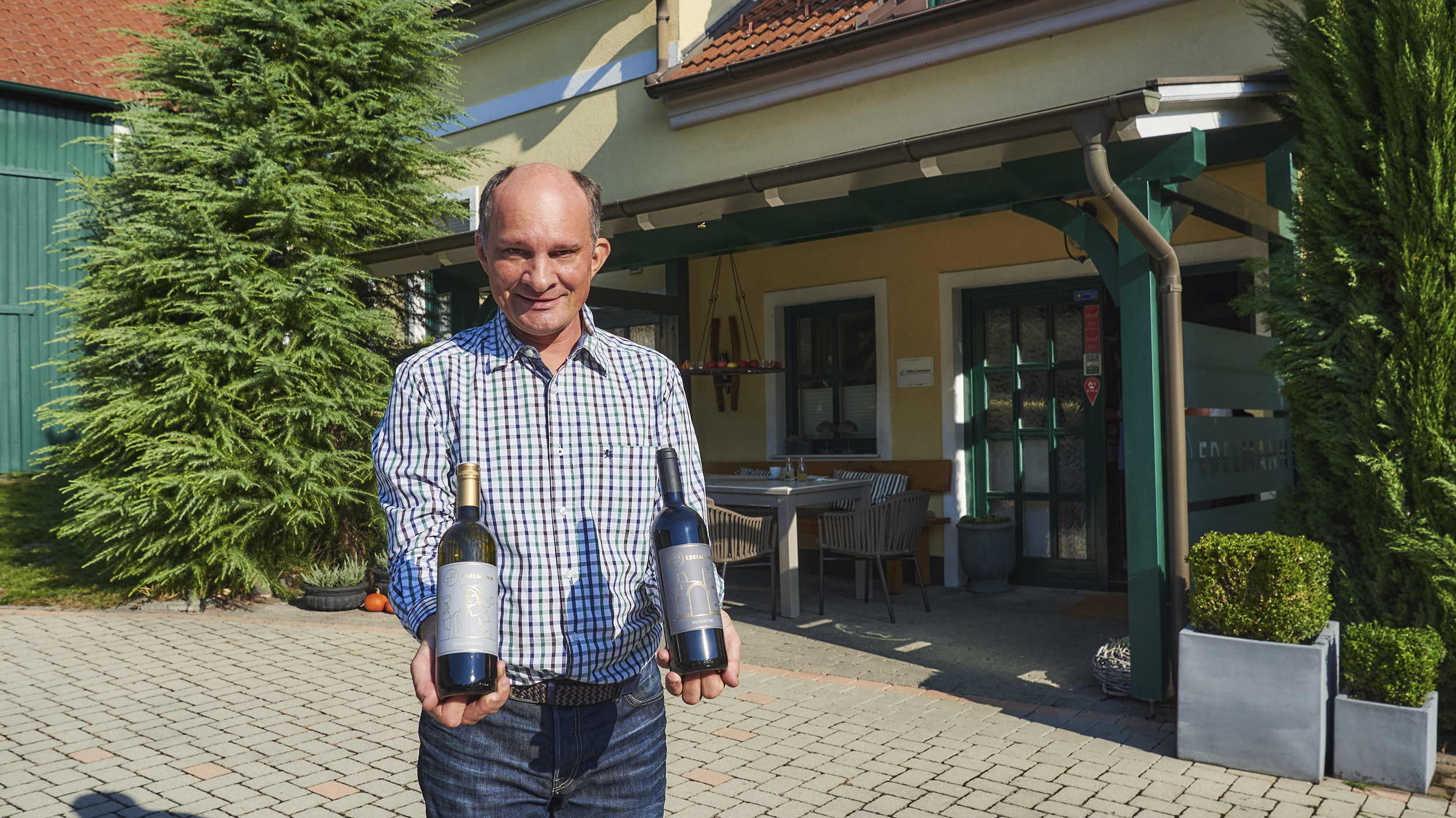 Winzer Manfred Edelmann präsentiert im Hof seines Betriebs zwei Weinflaschen, eine mit Chardonnay und eine mit einer Rotwein-Cuvée.