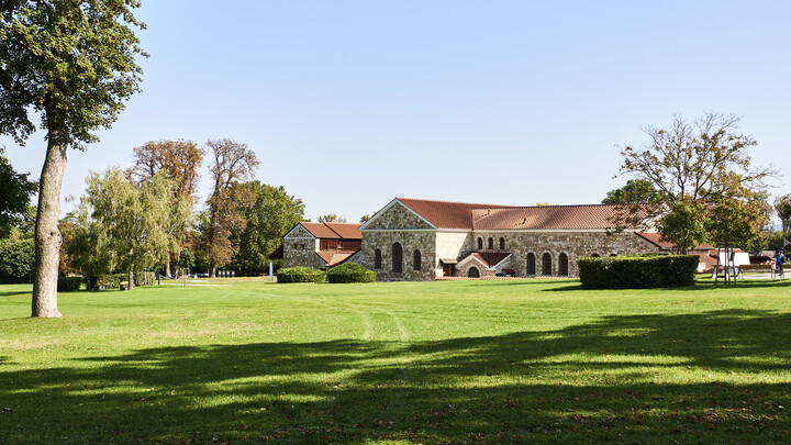 Hinter der grünen Wiese ist ein wieder aufgebautes großes römisches Haus aus Stein mit einem Ziegeldach zu sehen.