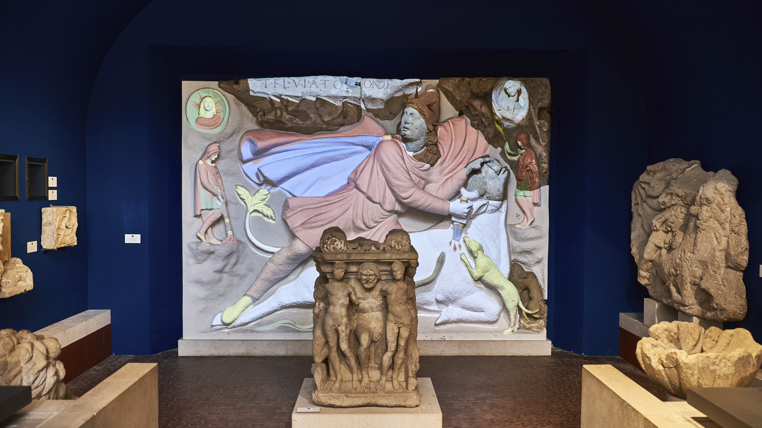 Figurengruppen aus Carnuntum und ein bemaltes Relief im Hintergrund.