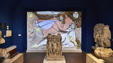 Figurengruppen aus Carnuntum und ein bemaltes Relief im Hintergrund.