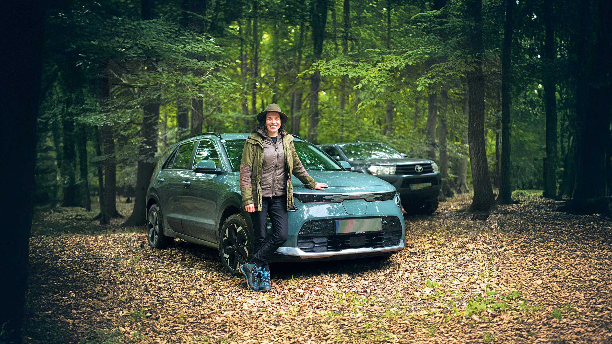 Eine Försterin lehnt an ihrem Auto, das mitten im Wald steht. Dahinter steht ein weiteres Fahrzeug.