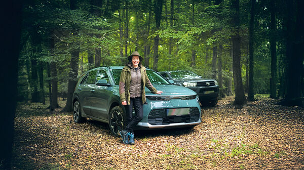 Eine Försterin lehnt an ihrem Auto, das mitten im Wald steht. Dahinter steht ein weiteres Fahrzeug.