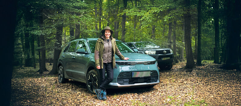 Eine Försterin lehnt an ihrem Auto, das mitten im Wald steht. Dahinter steht ein weiteres Fahrzeug.