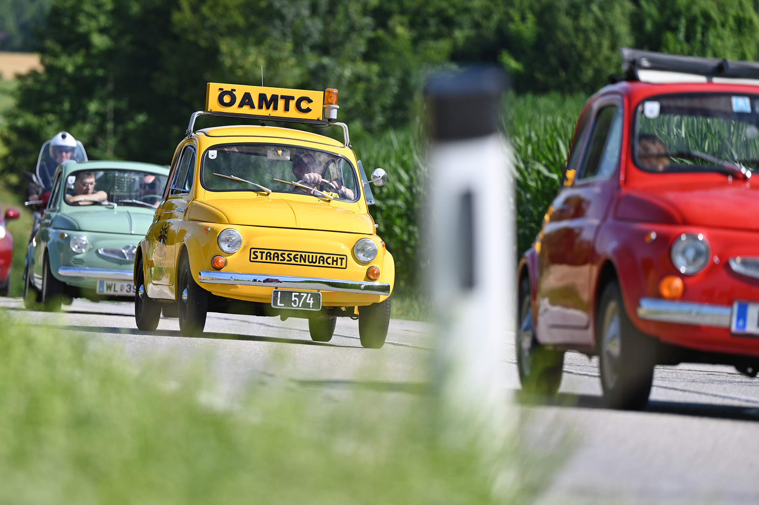 Kleine bunte Autos fahren auf einer Landstraße. Eines der Autos ist gelb und hat ein ÖAMTC-Schild auf dem Dach.