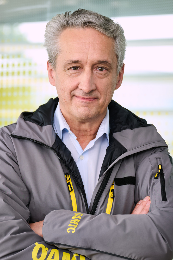Das Bild ist ein Portrait von Steffan Kerbl, der frontal in die Kamera schaut, eine graue Jacke mit gelber ÖAMTC-Aufschrift trägt und die Arme verschränkt.