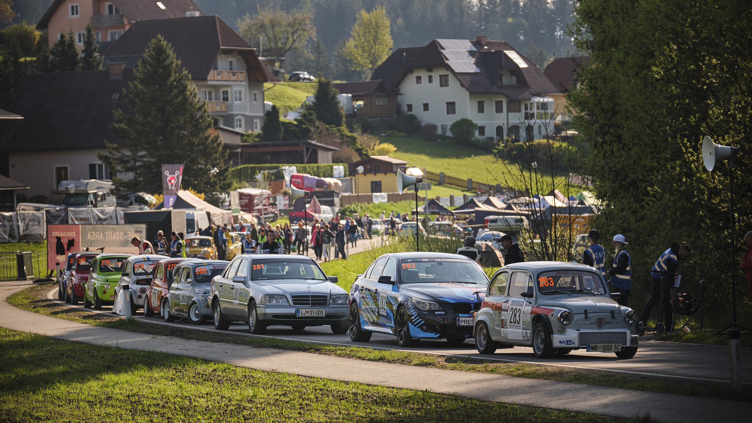 Rennautos stehen auf einer Landstraße, drumherum gehen Menschen.