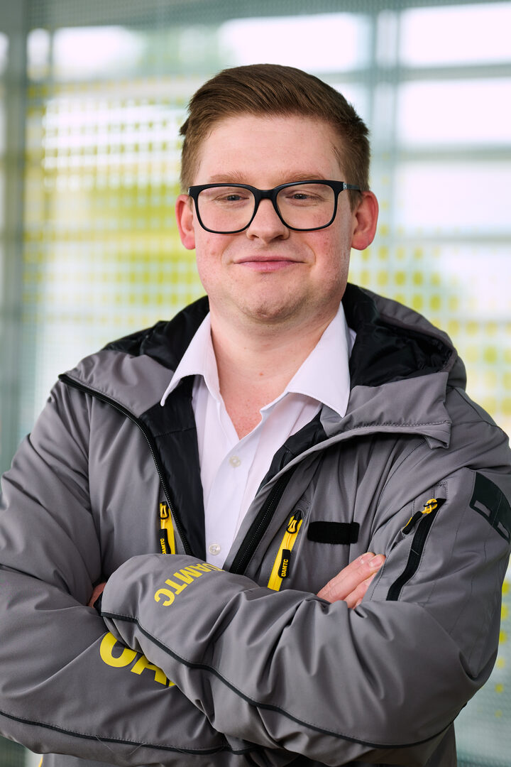 Porträt eines jungen Mannes mit kurzen rötlichen Haaren, einer Brille und verschränkten Armen in einer grau-gelben Jacke eines ÖAMTC-Technikers