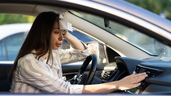 Auf dem Bild ist eine Frau im Auto zu sehen, die sich ein Tuch an die Stirn hält und mit der anderen Hand am Armaturenbrett legt. Der Frau ist sichtlich heiß und sie kontrolliert mit der Hand, ob die Klimaanlage funktioniert.