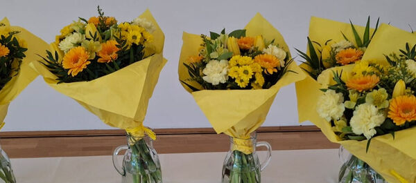 Das Foto zeigt fünf Blumensträuße mit gelbem Geschenkpapier umwickelt. Die Blumen selbst sind ebenfalls gelb und orange.