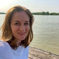 Das Bild ist ein Portrait von Barbara Gall. Sie hat schulterlange Haare und ein weißes T-Shirt an. Im Hintergrund ist ein See zu sehen.