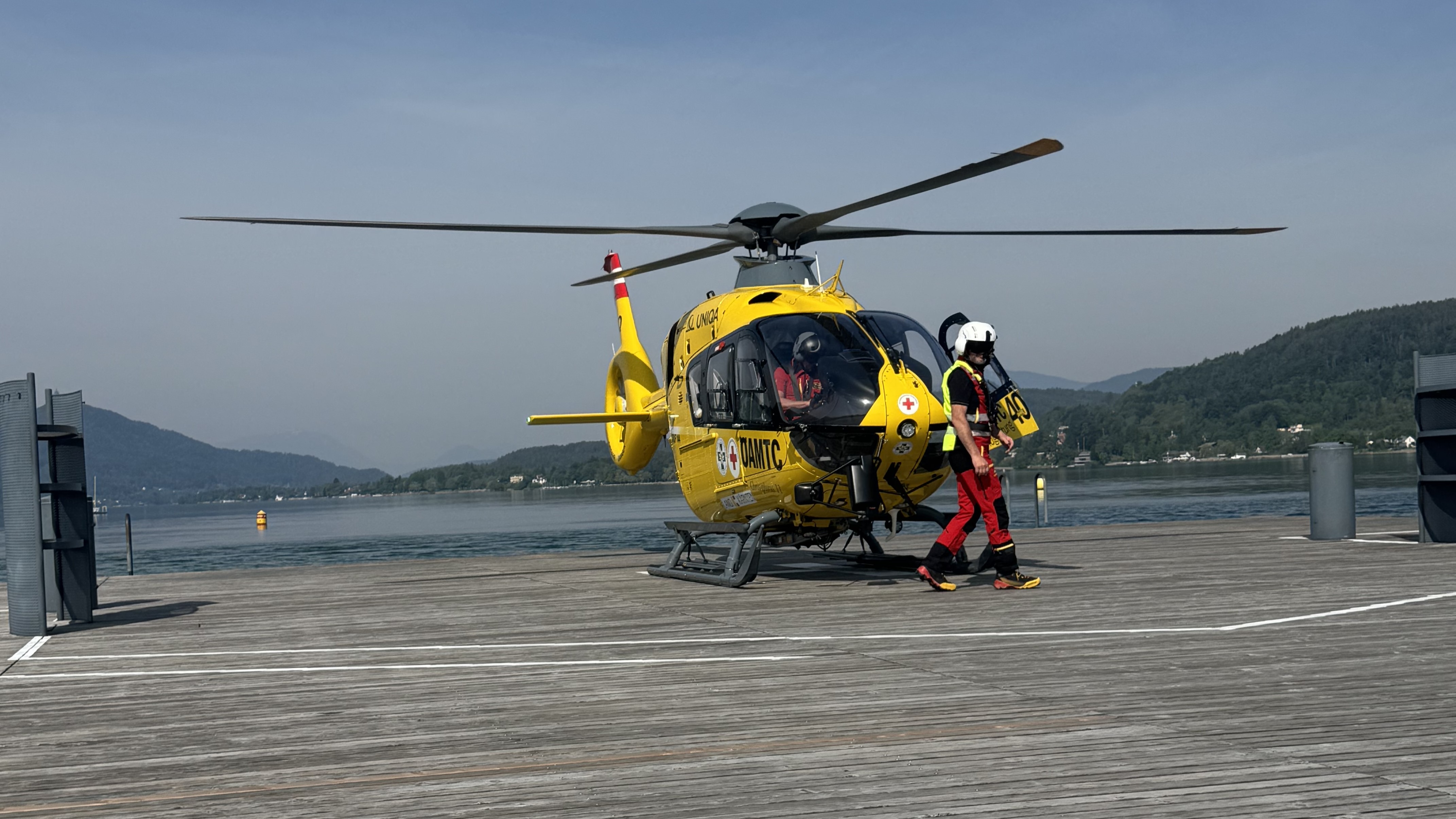 Christophorus-Hubschrauber steht auf einem Steg am See. Vor dem Hubschrauber geht ein Crew-Mitglied.