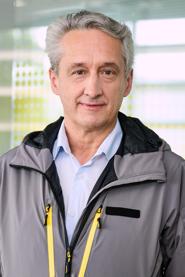 Das Foto zeigt einen Mann in einer grauen Jacke, unter der er ein hellblaues Hemd trägt. Der Mann hat graue Haare und steht vor einer Glaswand mit gelben Punkten.