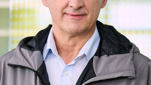 Das Foto zeigt einen Mann in einer grauen Jacke, unter der er ein hellblaues Hemd trägt. Der Mann hat graue Haare und steht vor einer Glaswand mit gelben Punkten.