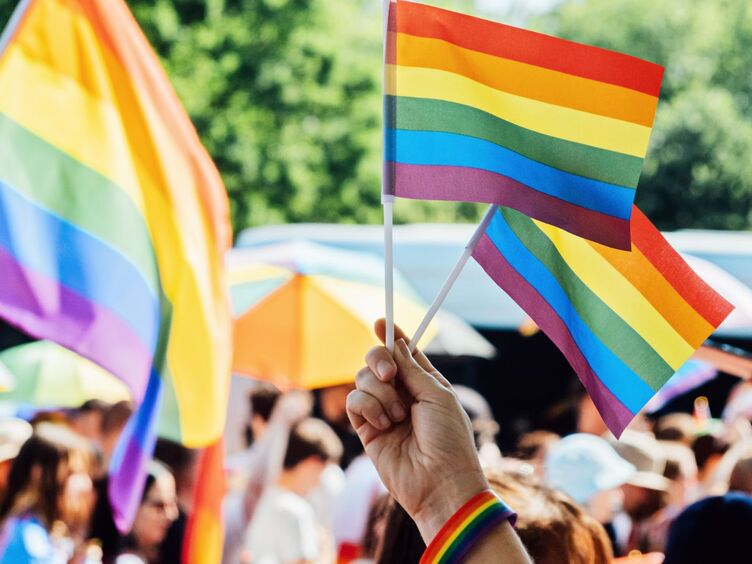 Header Blog Queer Angels Auf dem Foto sind viele Regenbogenfahnen abgebildet. Im Vordergrund sieht man nur eine Hand, die ein Regenbogenarmband trägt und zwei kleine Regenbogenfahnen in die Luft hält.