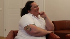 Ursula im Interview über Queersein im Arbeitsalltag. Das Bild zeigt eine Frau von der Seite. Sie sitzt und stütz einen Arm am Kinn ab, sie trägt eine Brille und ein weißes Polo-Shirt.