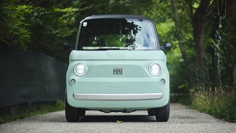 Ein türkises Microcar steht auf einer einspurigen Straße, die von Bäumen umgeben ist.
