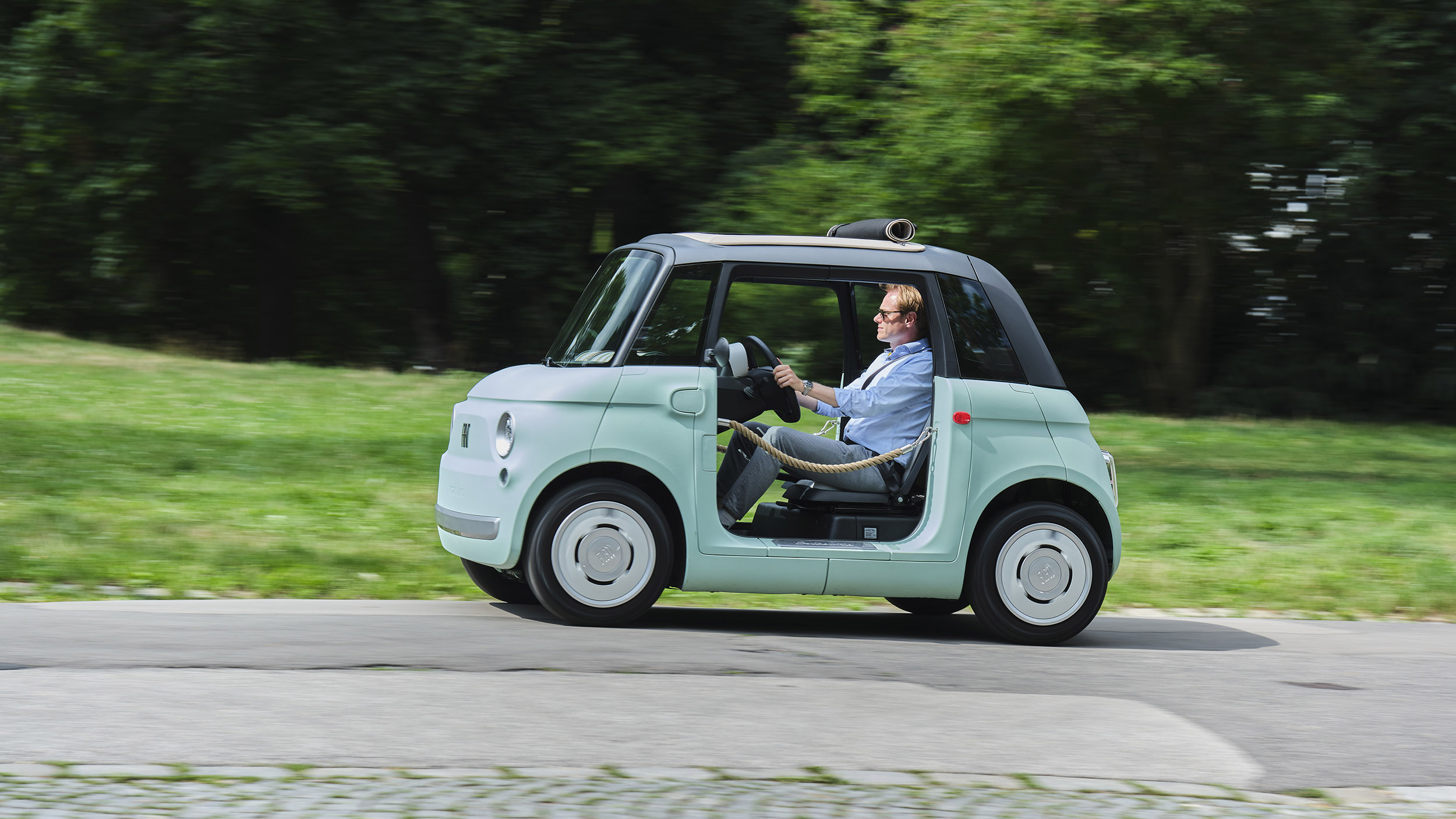 Eine Person fährt mit einem türkis-schwarzen Microcar, das anstelle von Türen braune Flechtkordeln hat, über eine Straße, die an einer grünen Wiese und Bäumen entlang führt.