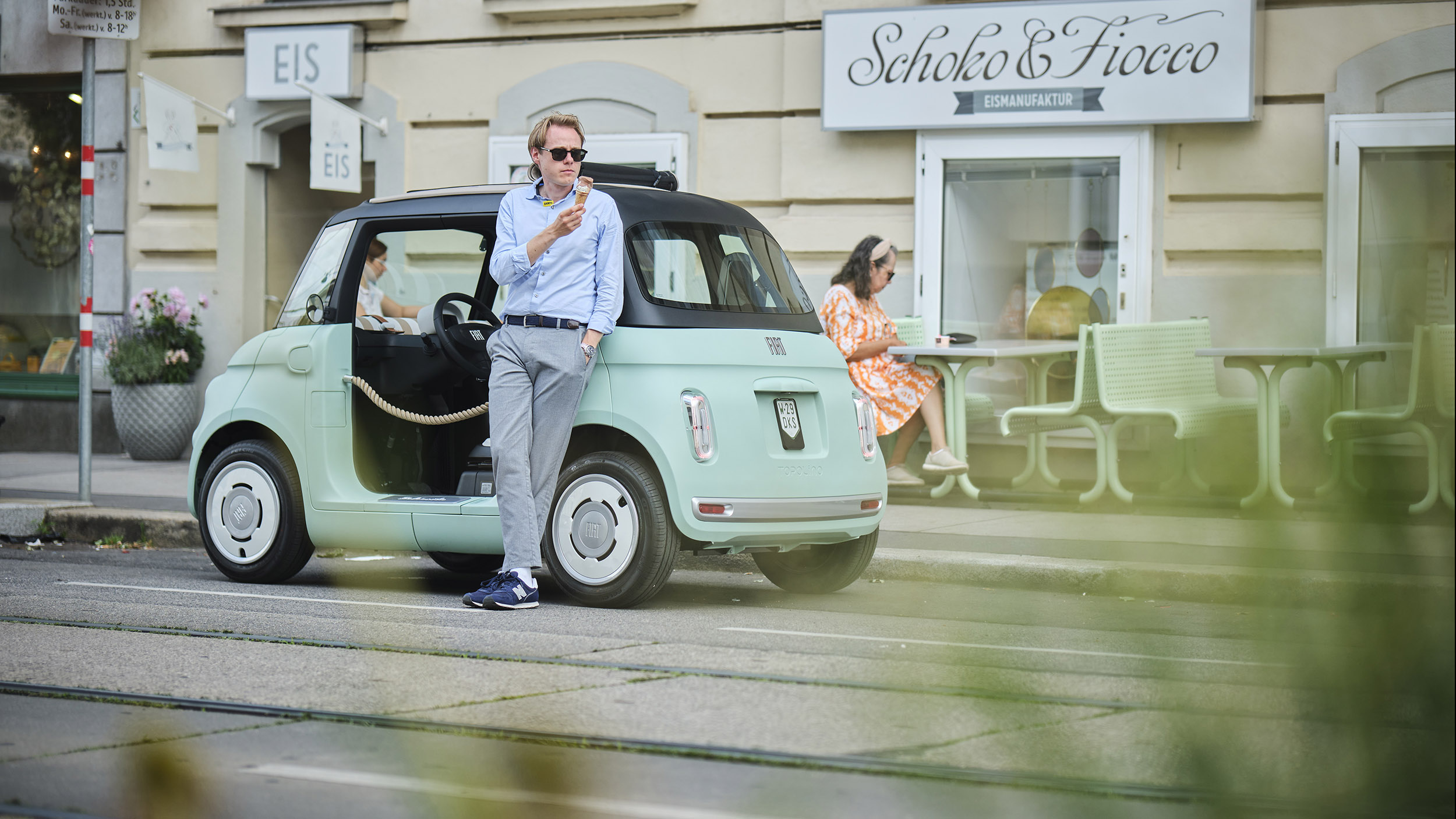Eine Person in blauem Hemd und grauer Hose lehnt an einem türkisen Microcar, das vor einem Eissalon in der Stadt parkt, und isst Eis.
