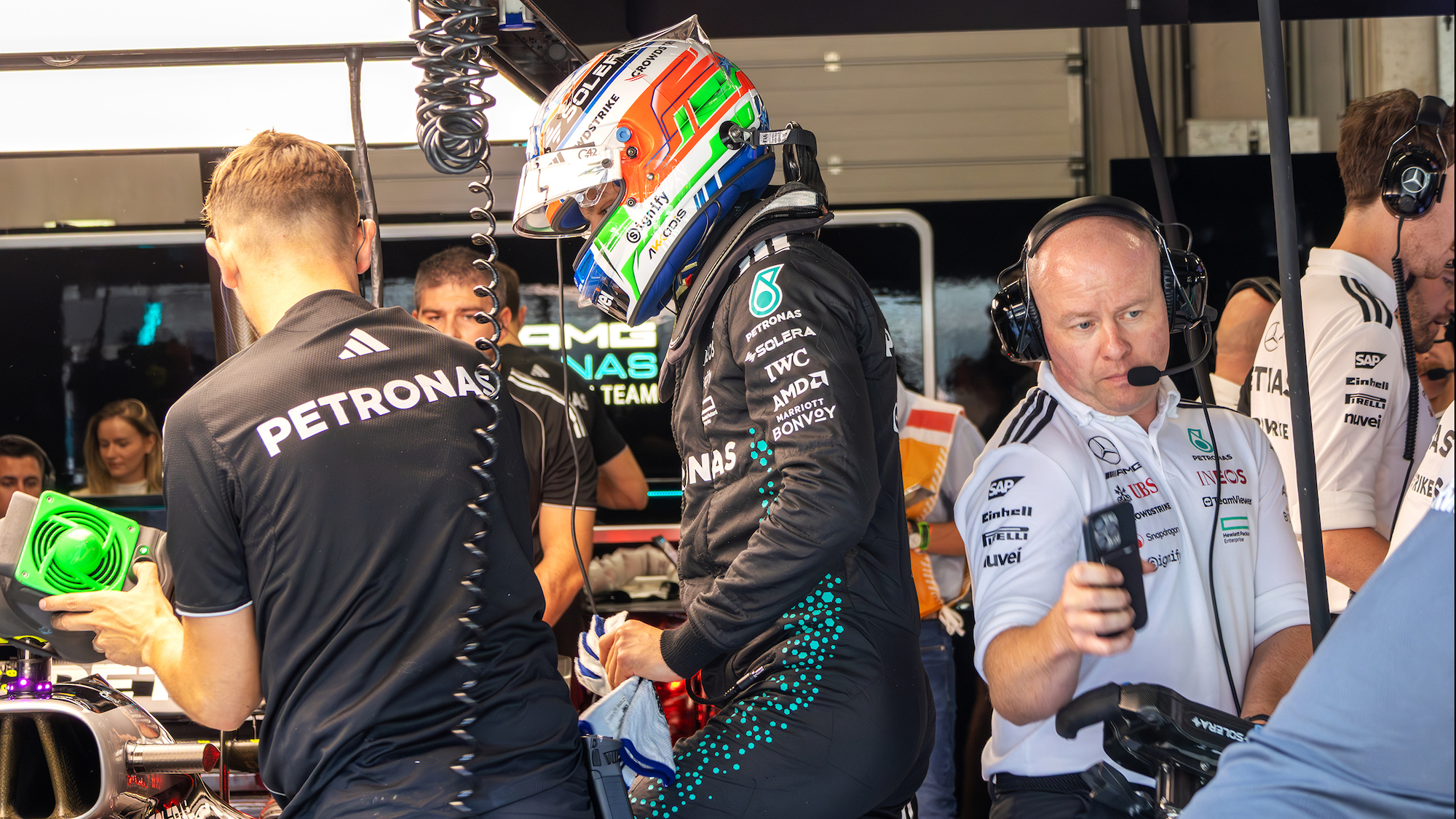 Ein Formel-1-Fahrer in voller Montur mit Rennanzug und -helm steht neben seinem Formel 1 Auto, um ihn herum arbeiten viele Teammitglieder.