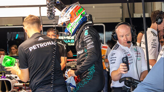 Ein Formel-1-Fahrer in voller Montur mit Rennanzug und -helm steht neben seinem Formel 1 Auto, um ihn herum arbeiten viele Teammitglieder.