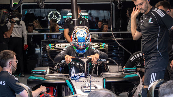 Ein Formel-1-Fahrer in voller Montur mit Rennanzug und -helm steht neben seinem Formel 1 Auto, um ihn herum arbeiten viele Teammitglieder.