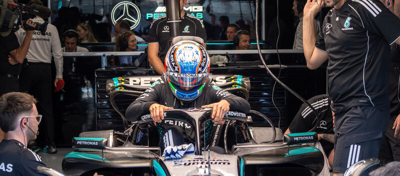 Ein Formel-1-Fahrer in voller Montur mit Rennanzug und -helm steht neben seinem Formel 1 Auto, um ihn herum arbeiten viele Teammitglieder.