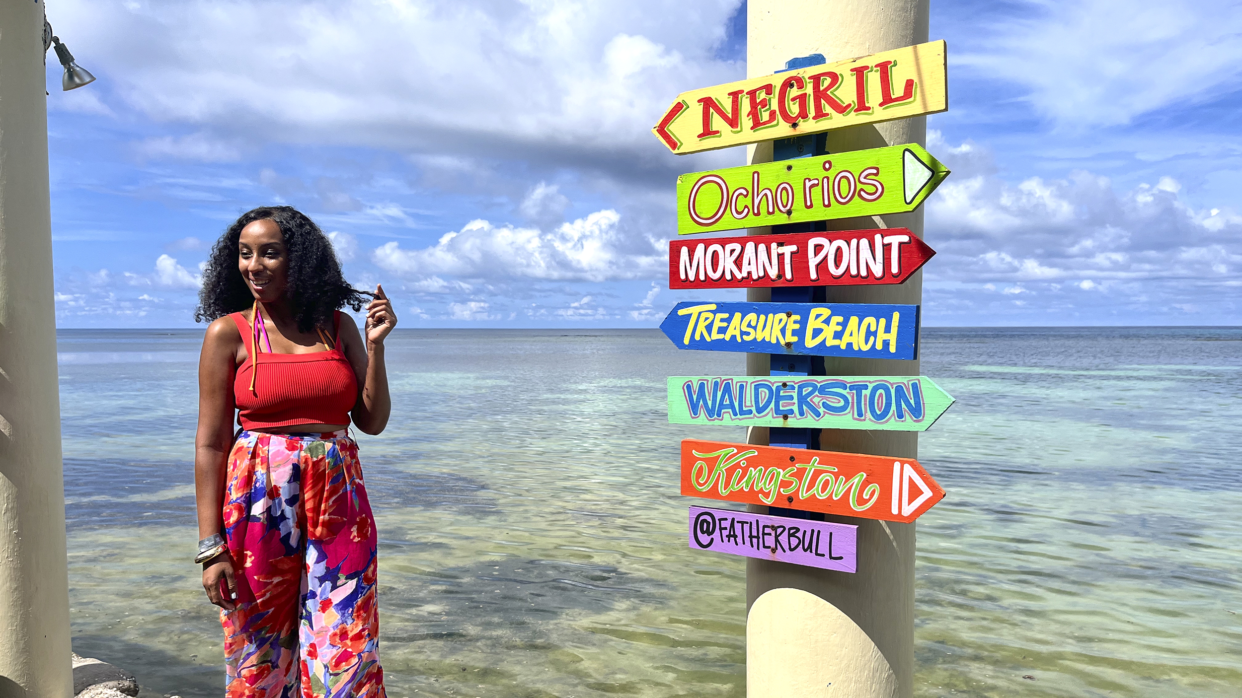 Bunte Ortsschilder am Meer und eine Dame steht daneben.