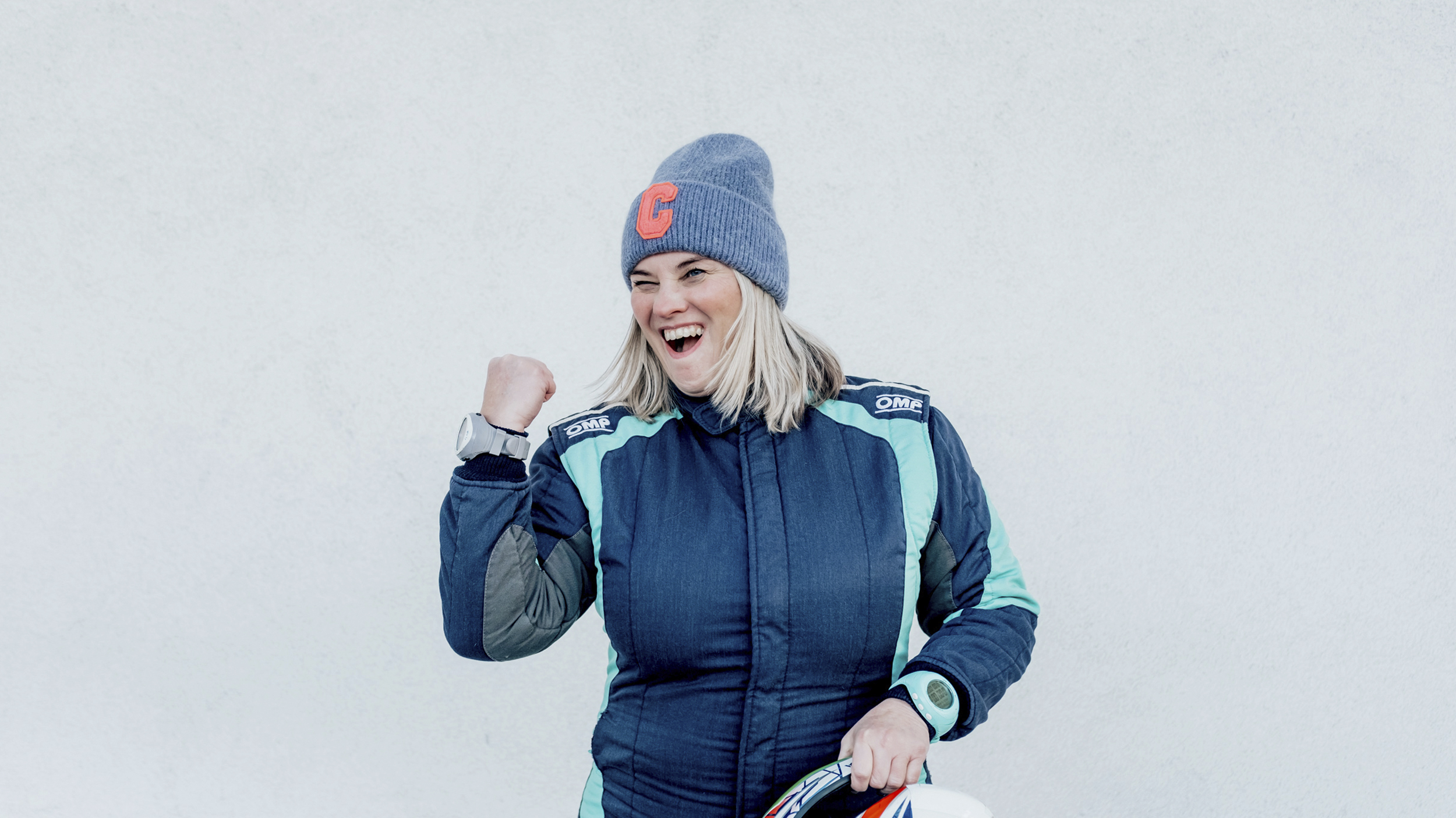 Catharina Schmidt, Rallye-Co-Pilotin und ÖAMTC-Mitarbeitetin steht im Renndress vor hellgrauem Hintergrund in Siegespose und mit Haube.