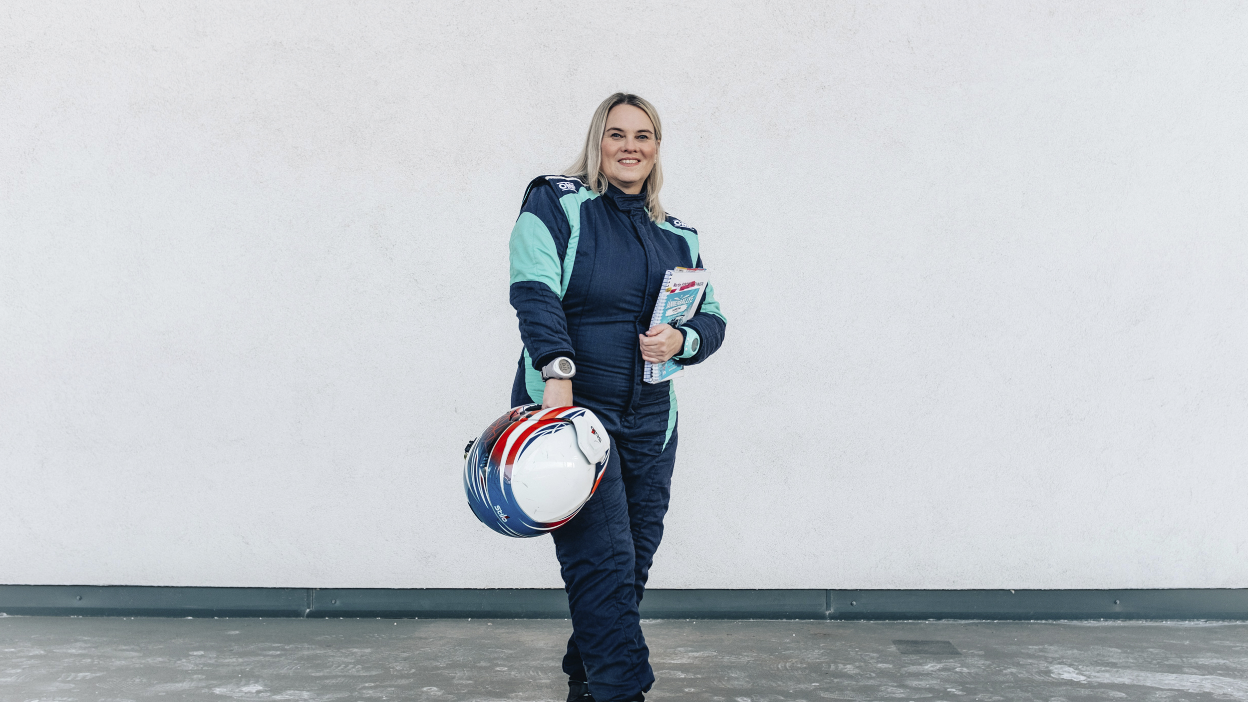 Catharina Schmidt, Rallye-Co-Pilotin und ÖAMTC-Mitarbeitetin steht im Renndress und mit Haube vor hellgrauem Hintergrund und Helm in der Hand.