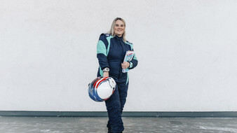 Catharina Schmidt, Rallye-Co-Pilotin und ÖAMTC-Mitarbeitetin steht im Renndress und mit Haube vor hellgrauem Hintergrund und Helm in der Hand.