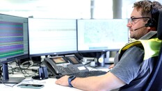 Auf dem Bild sieht man einen Mann mit Bart, der ein headset am Kopf hat und vor drei Computerbildschirmen sitzt.