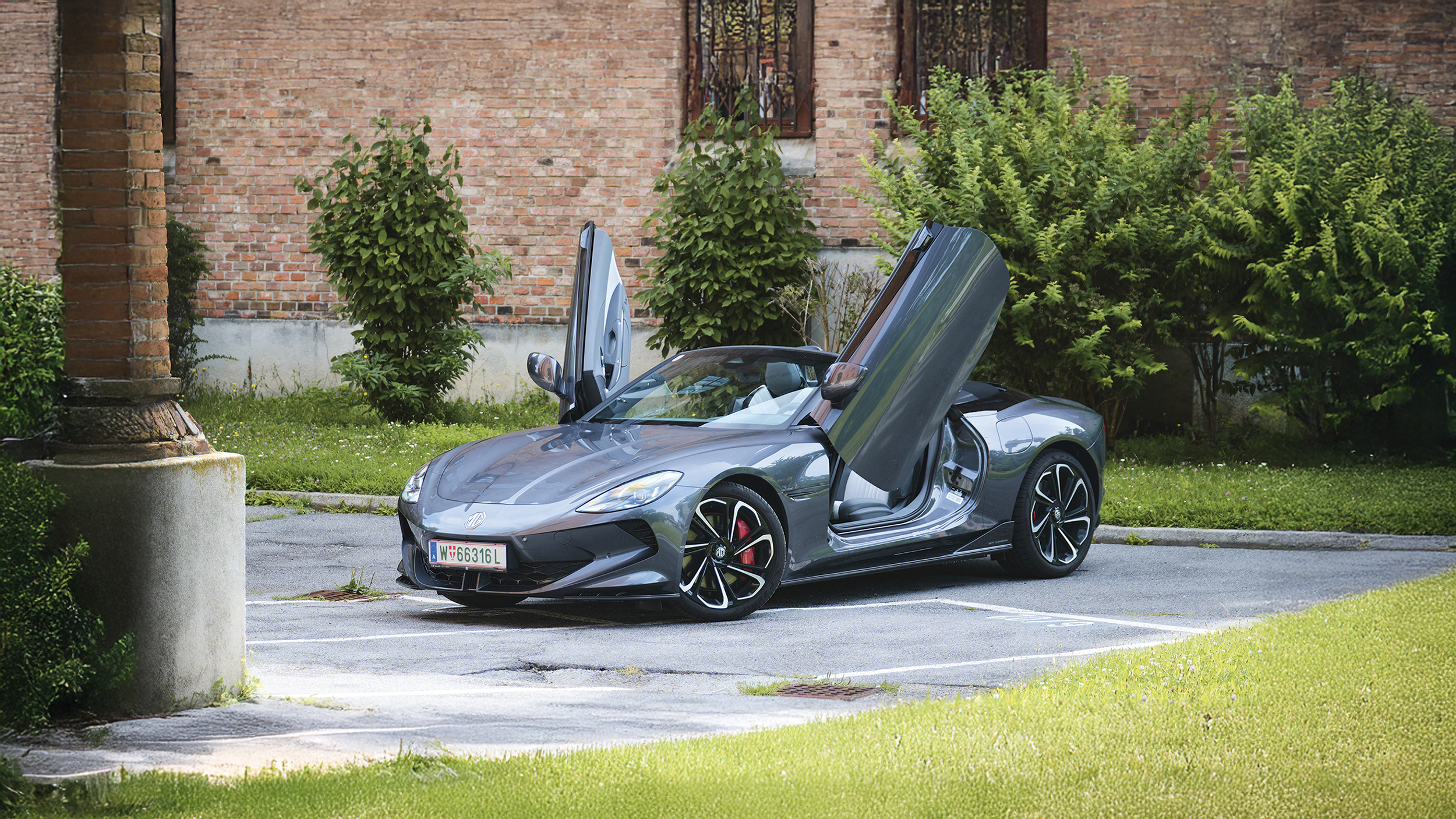 Ein graues Cabrio steht mit offenem Verdeck und Flügeltüren vor einem Backstein-Gebäude.