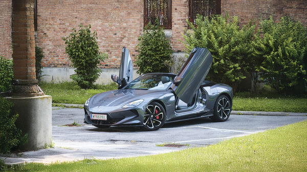 Ein graues Cabrio steht mit offenem Verdeck und Flügeltüren vor einem Backstein-Gebäude.