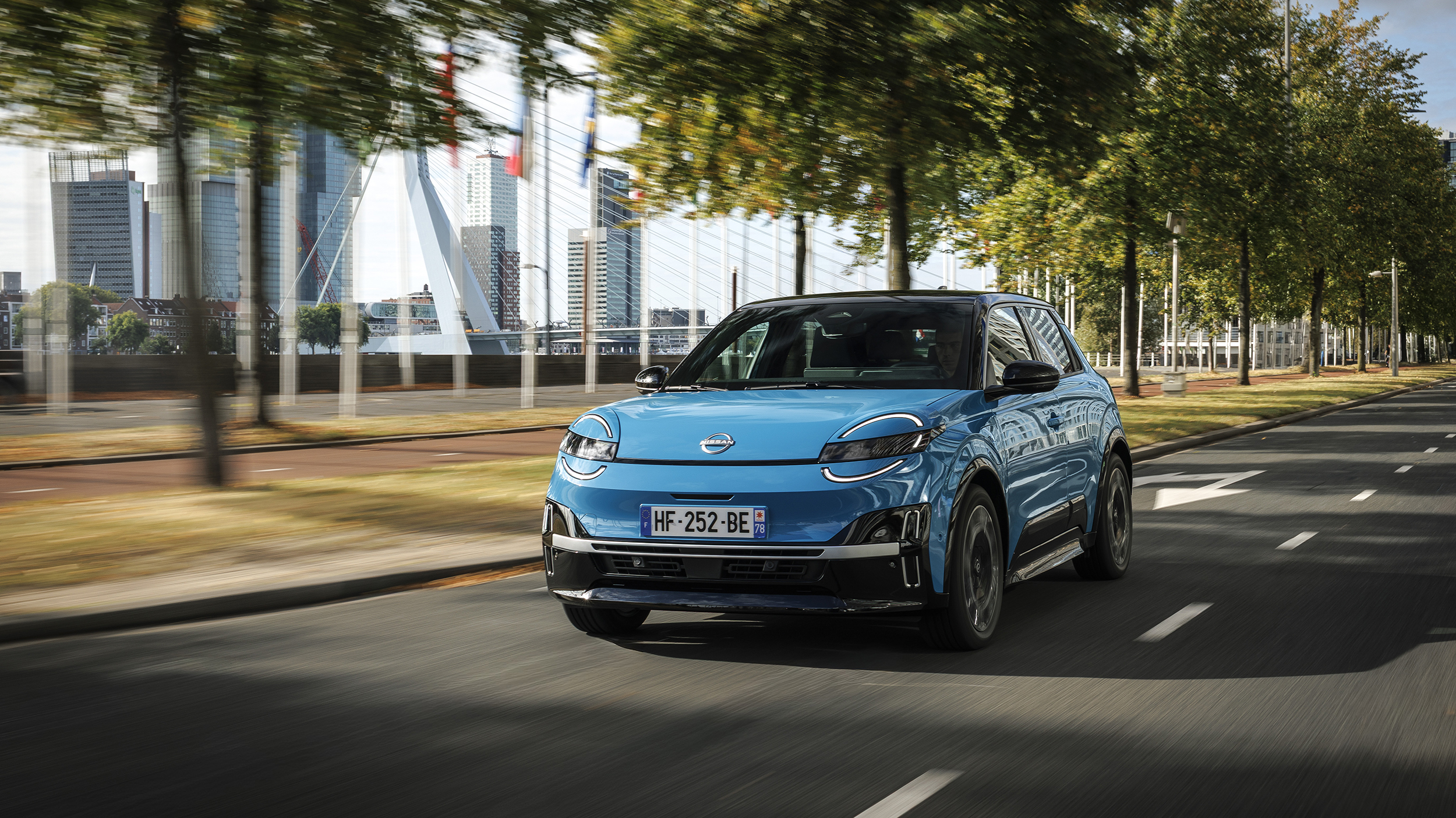 Ein blauer Nissan Micra 2025 fährt auf einer Straße, hinter der sich Bäumen und moderne Gebäude befinden.