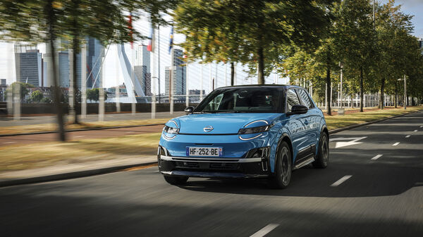Ein blauer Nissan Micra 2025 fährt auf einer Straße, hinter der sich Bäumen und moderne Gebäude befinden.