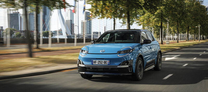 Ein blauer Nissan Micra 2025 fährt auf einer Straße, hinter der sich Bäumen und moderne Gebäude befinden.