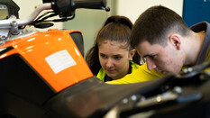 Auf dem Bild sieht man eine junge Frau und einen jungen Mann die konzentriert an einem Orangefarbenen Motorrad arbeiten.