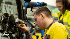 Auf dem Bild sieht man einen jungen Mann der ein Fahrrad repariert. Im Hintergrund beobachtet ihn eine Kollegin