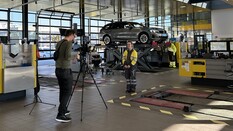 Auf dem Bild stehen ein Kameramann und eine Interviepartnerin in einer Werkstatt des ÖAMTC. Im Hintergrund sieht man einige Fahrzeuge.