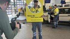 Auf dem Bild sieht man einen Kameramann, der in einer Werkstatt gerade inen ÖAMTC-Techniker fotografiert. Der Techniker hält einen gelben Rahmen in der Hans. Auf diesem kann man lesen: Gönn dir einen neuen Job