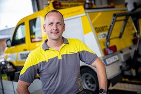 Das Bild ist ein Portraitfoto des Pannenfahrers Andi, er trägt ein gelb-graues ÖAMTC-Shirt und im Hintergrund ist unscharf ein ÖAMTC-Pannenfahrzeug.